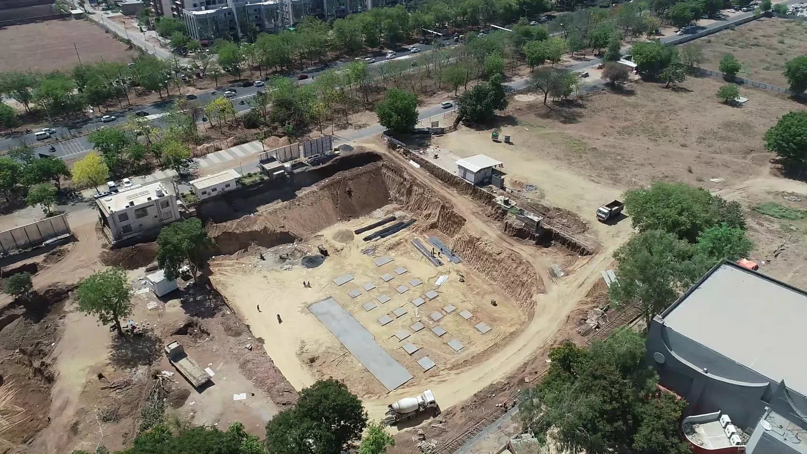 Aerial view of an excavated building site with foundation pads, trucks and trees along the road