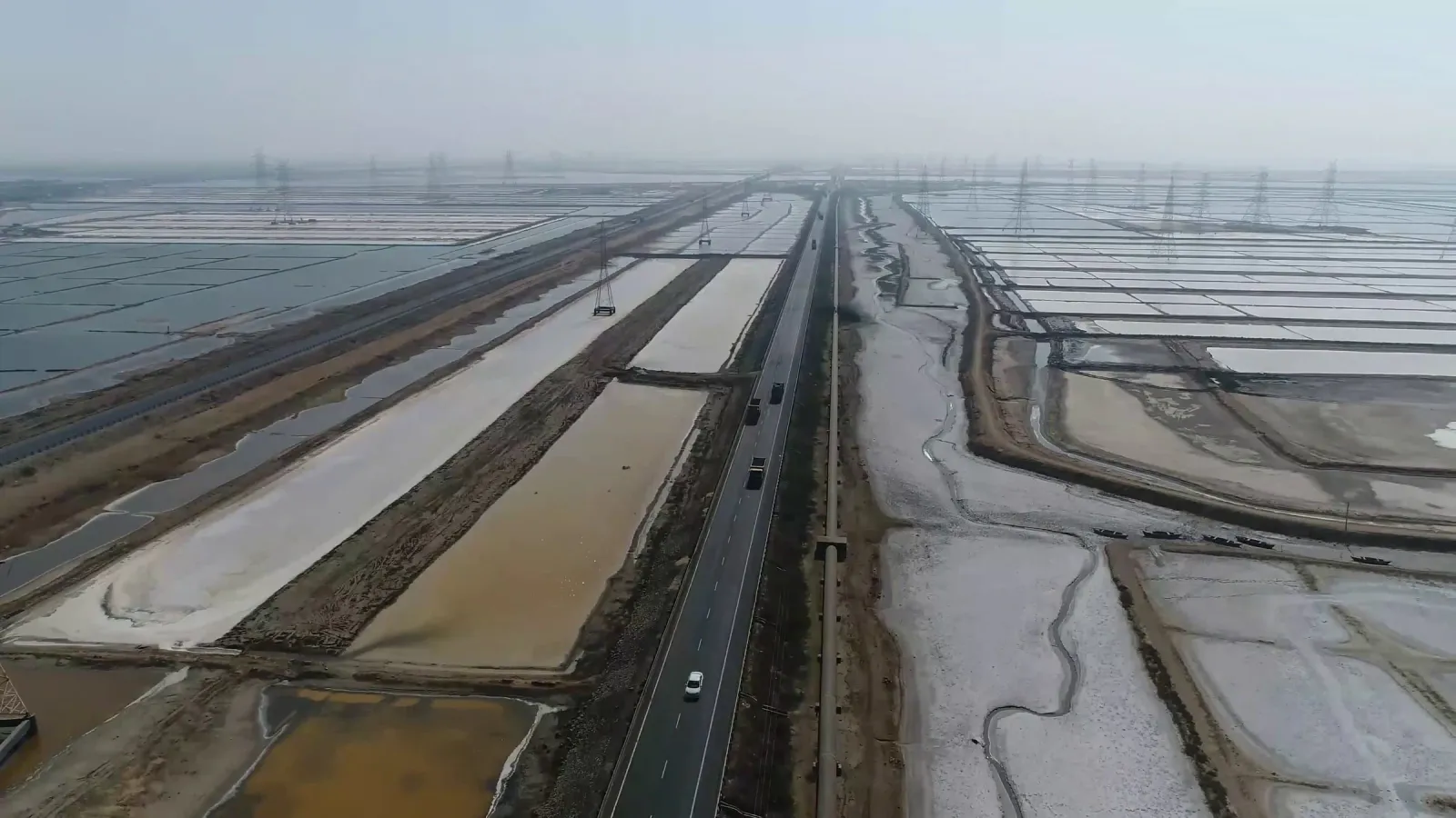 Aerial view along a highway cutting through flooded salt pans under a hazy sky