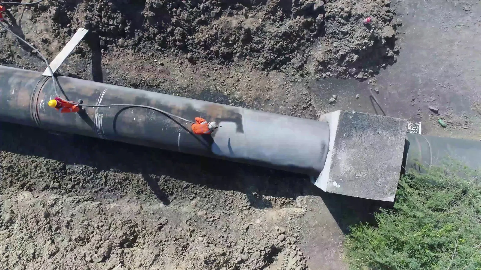 Aerial view straight down on two workers in orange on a large steel pipeline beside an earth trench