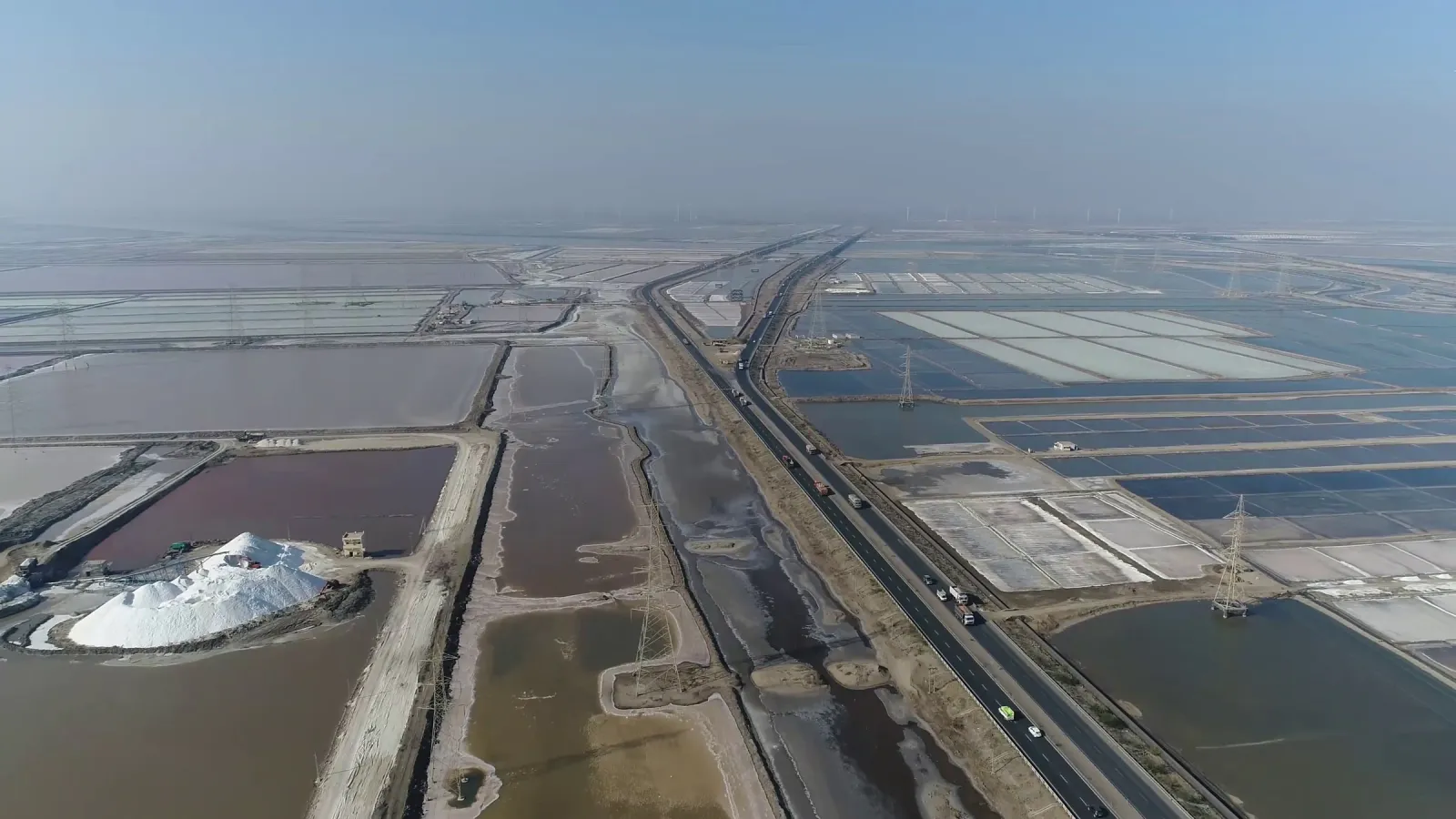 Aerial view of salt pans with a white salt heap beside a highway running towards the horizon