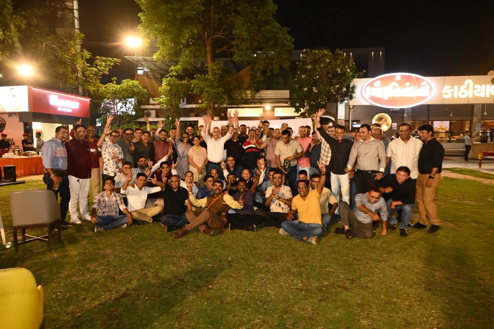 Large cheerful group posing on a lawn at a night-time gathering outside a restaurant