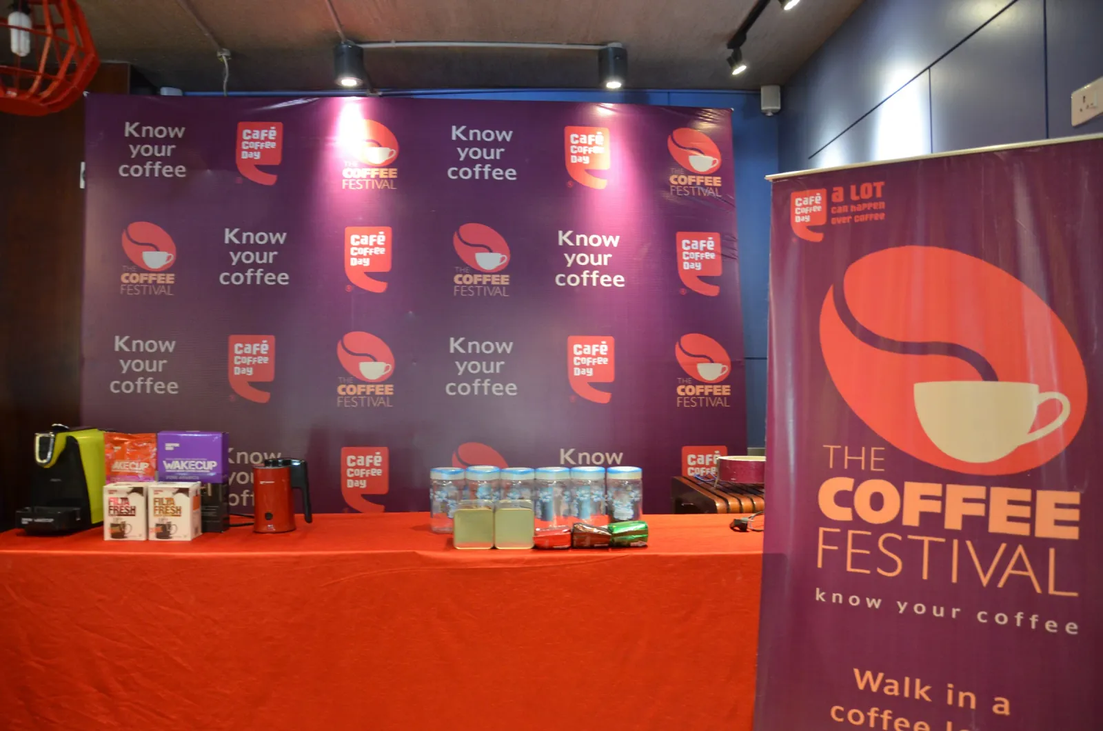 Coffee festival display table with coffee machines, cups and product boxes in front of a purple Know Your Coffee backdrop