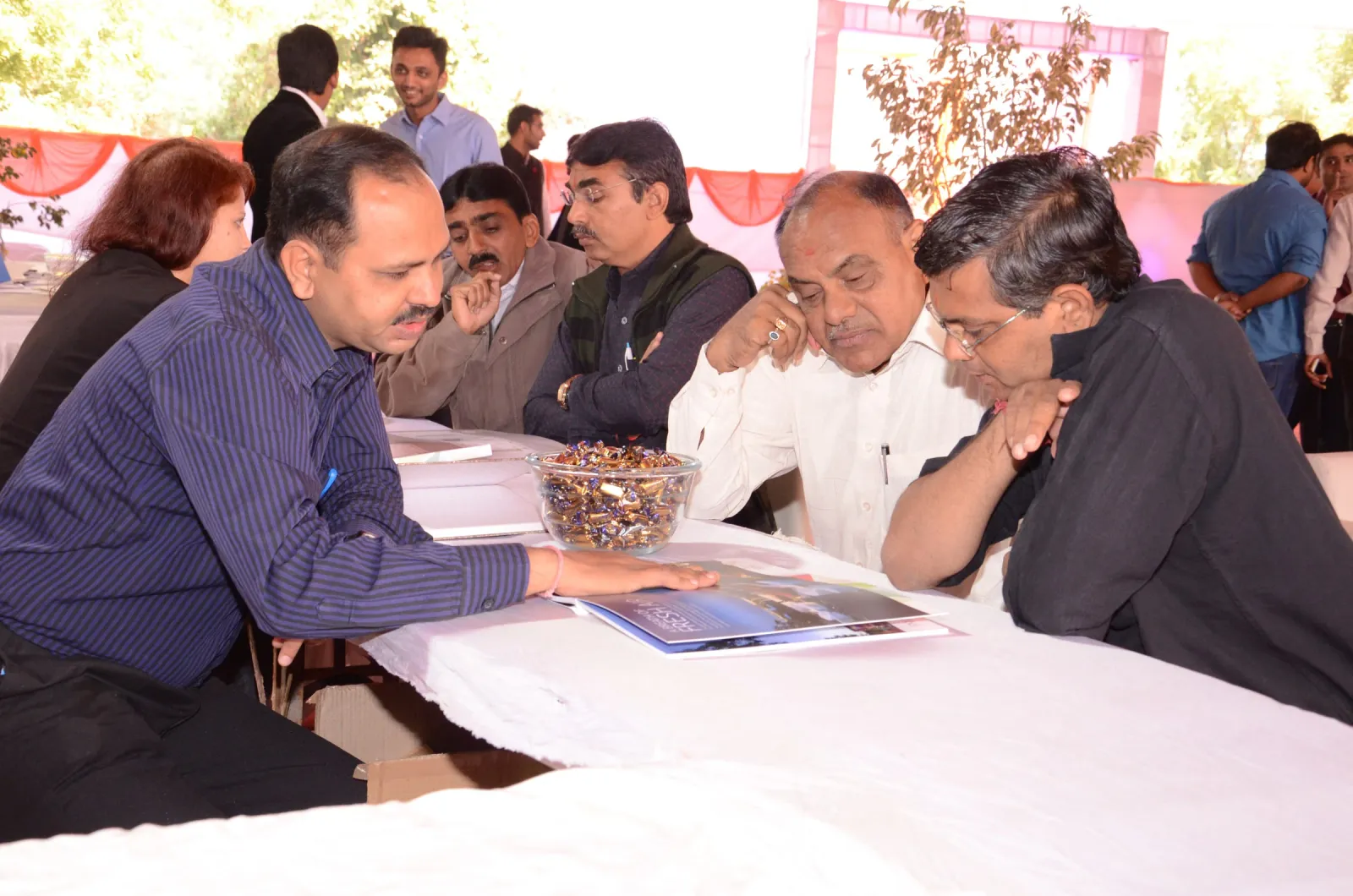 Men seated at a white-clothed table reading a brochure together at an outdoor event under a tent