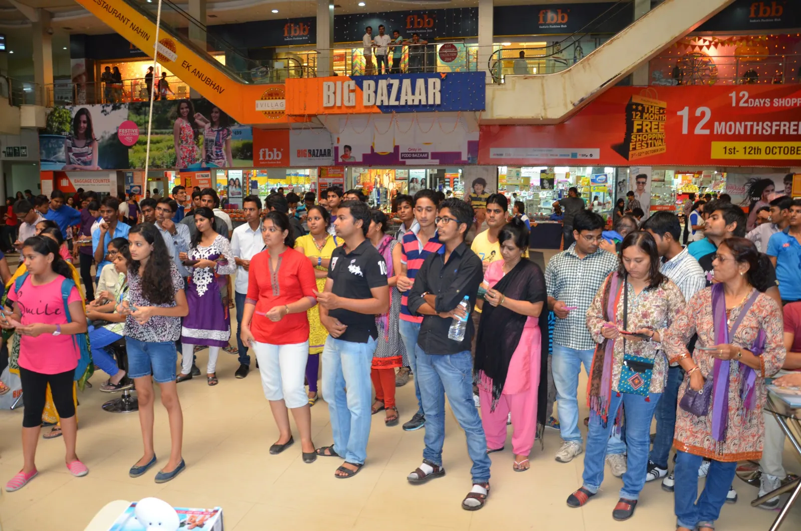 Shoppers standing in a mall atrium watching an event, with a Big Bazaar storefront behind them