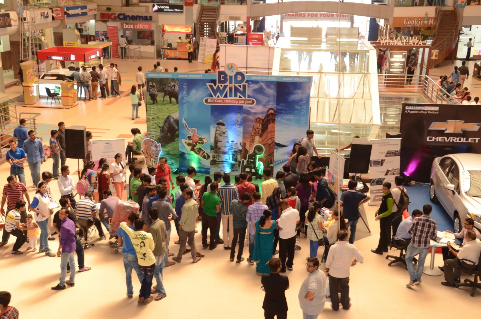 Crowd gathered around a Bid and Win promotion stage and a car display in a shopping mall atrium, seen from the floor above