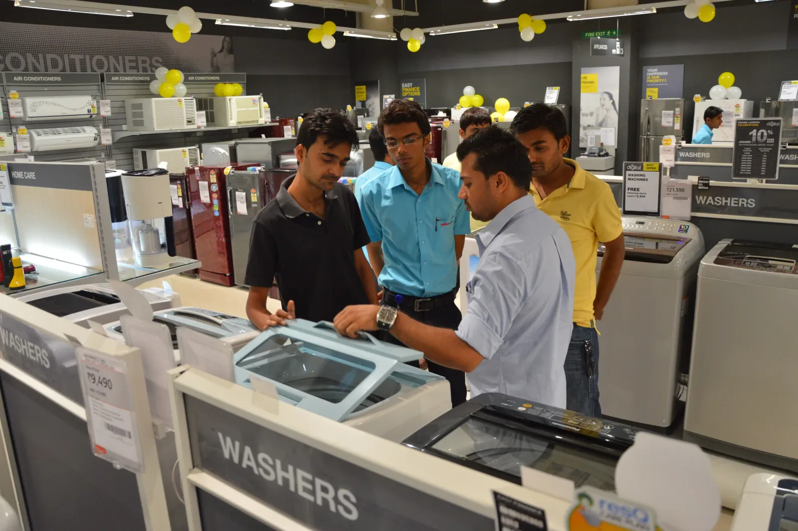 Staff member showing a top-loading washing machine to three customers in the appliance section of an electronics store