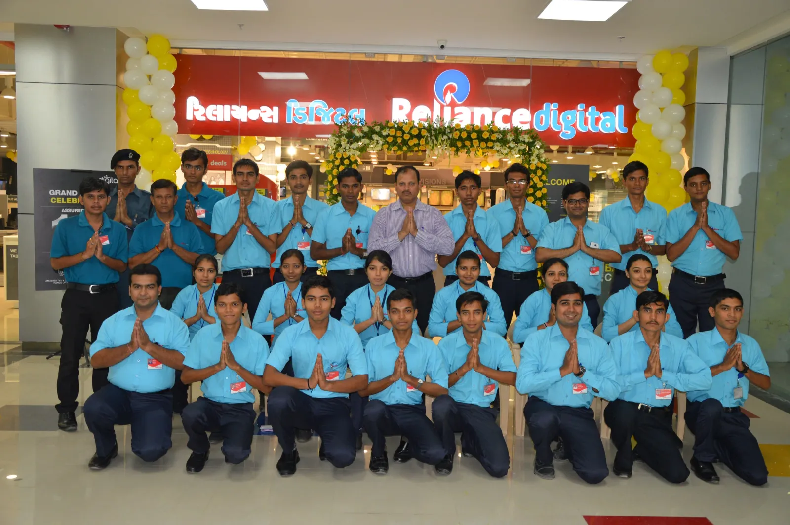 Store staff in blue uniforms kneeling and standing with folded hands under the Reliance Digital sign at the entrance