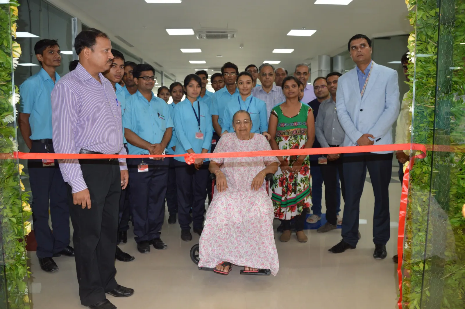 Elderly woman in a wheelchair cutting the red ribbon at a store opening, with staff in blue shirts and guests standing.