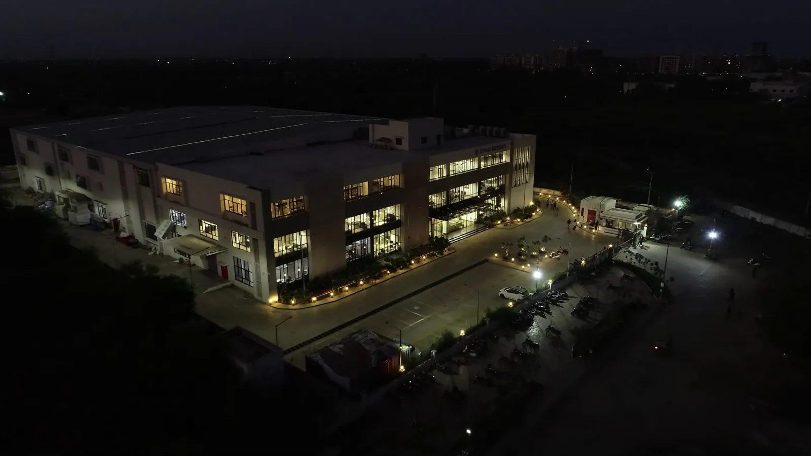Aerial view at dusk of a lit three-storey office building with a parking lot and a warehouse behind it