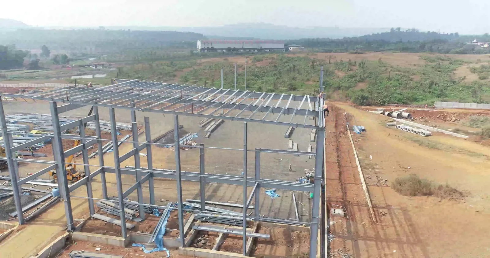 Aerial view of a steel-framed factory shed under construction on red soil with hills in the distance