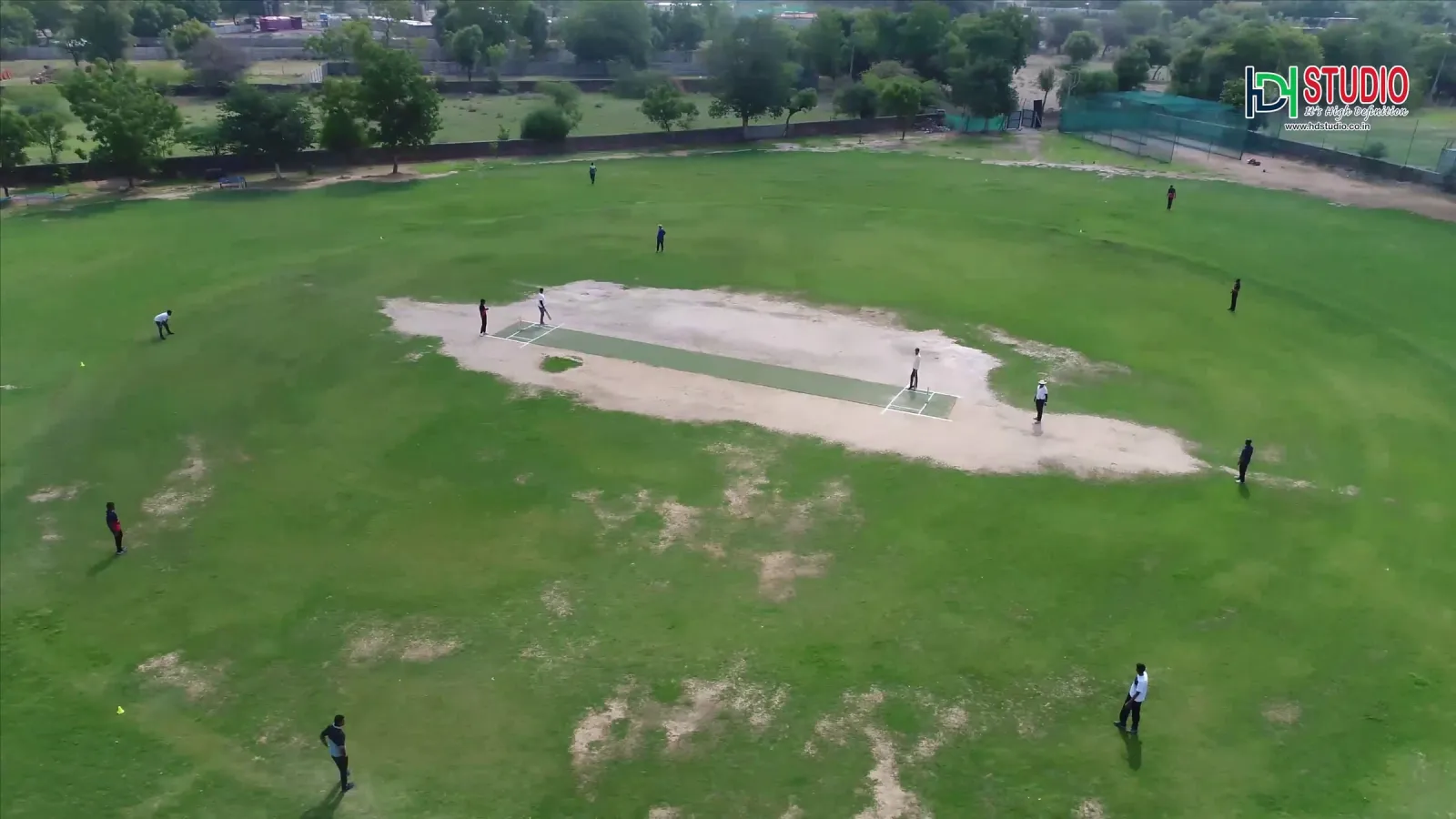 Aerial view of a cricket match in progress on a green ground with the pitch in the centre and fielders spread out