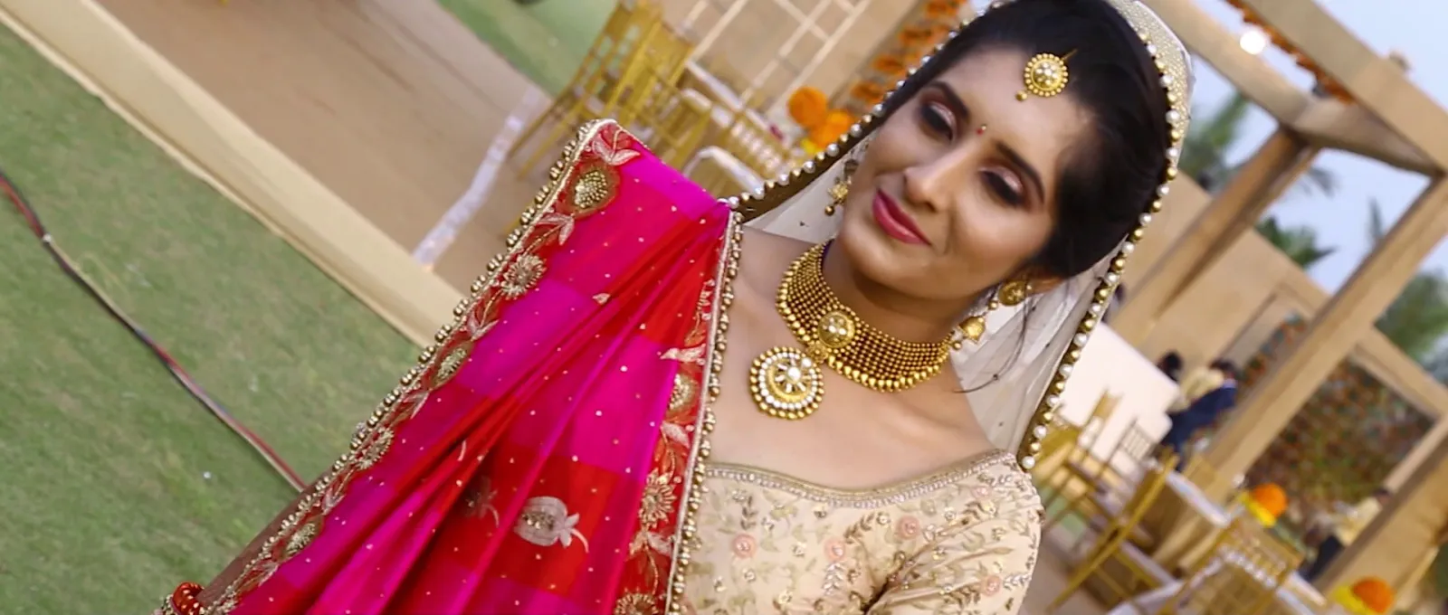 Bride in a pink and gold dupatta with gold jewellery looking to the side at an outdoor wedding venue