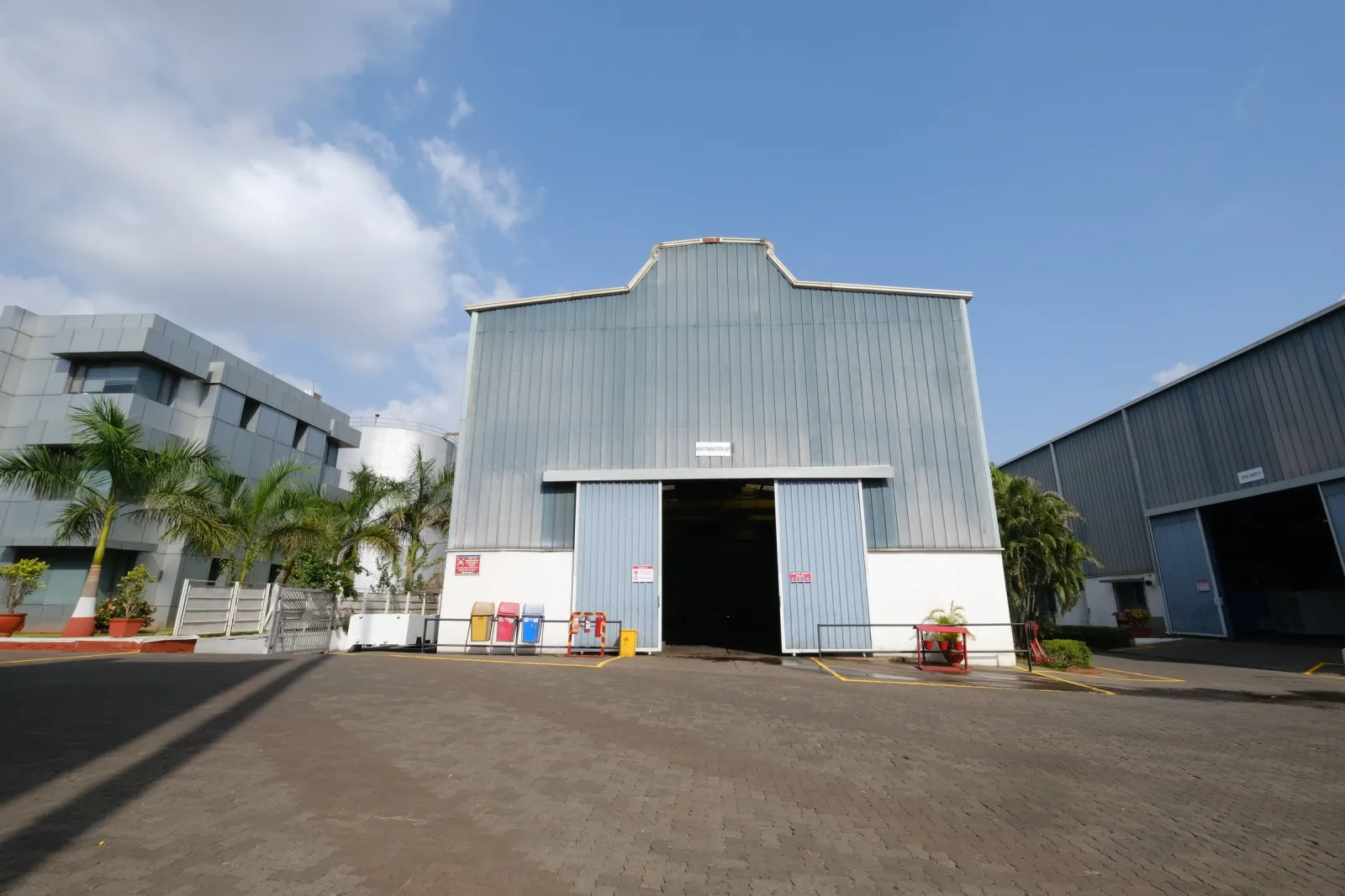 Heavy fabrication shed with open sliding doors, palm trees and an office block under a blue sky