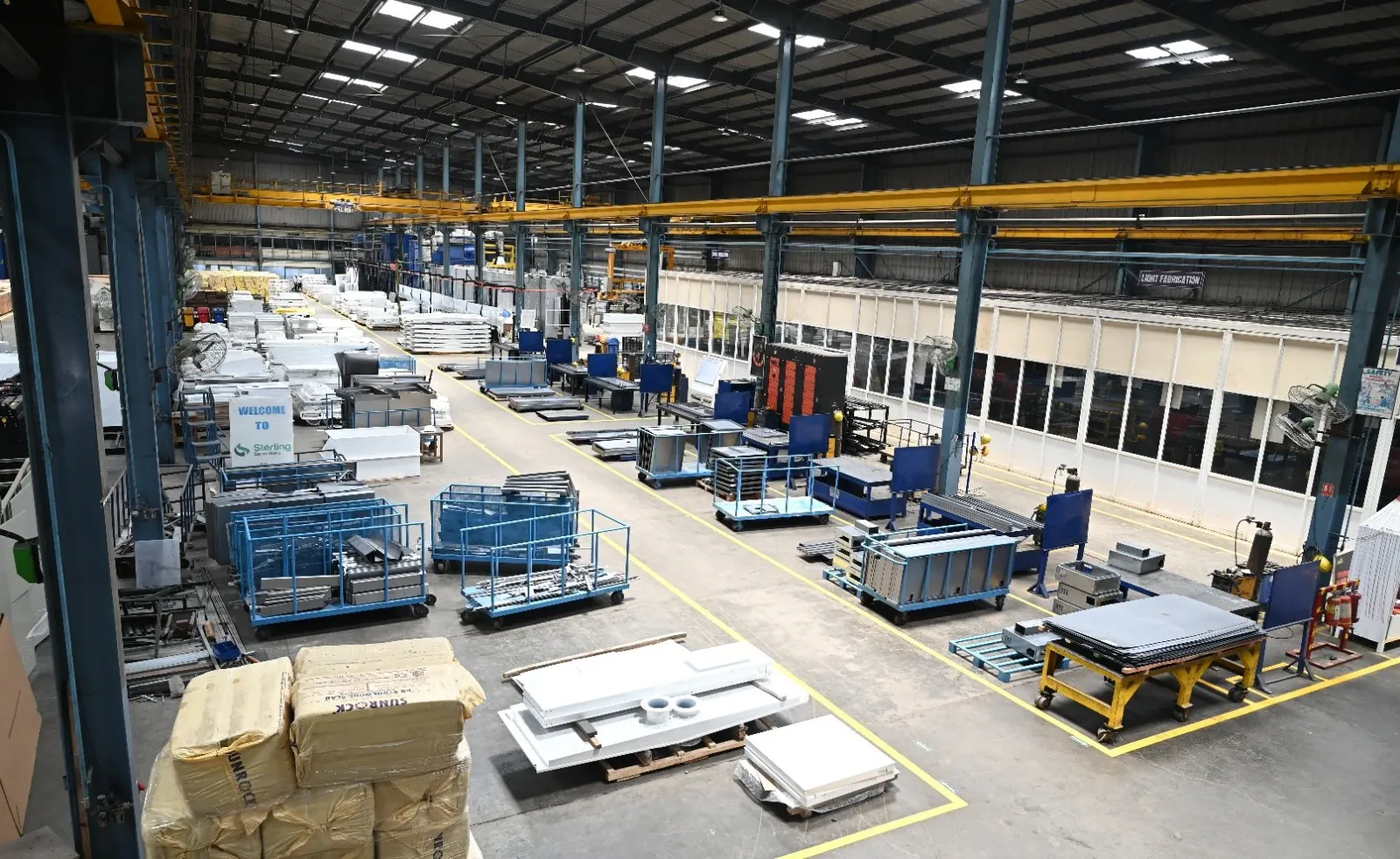 Wide view of a fabrication shop floor with overhead cranes, blue trolleys and sheet metal parts
