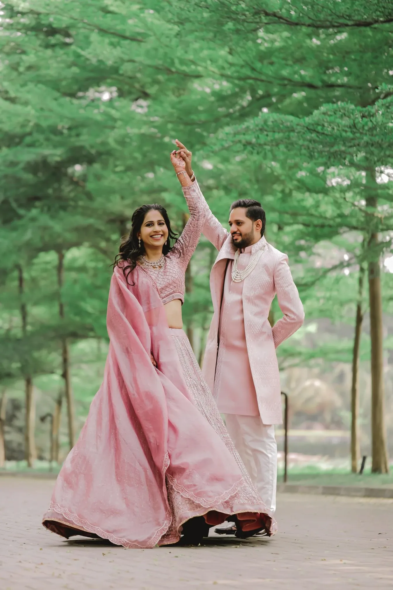 Couple in pink traditional outfits dancing on a tree-lined path during a pre-wedding shoot