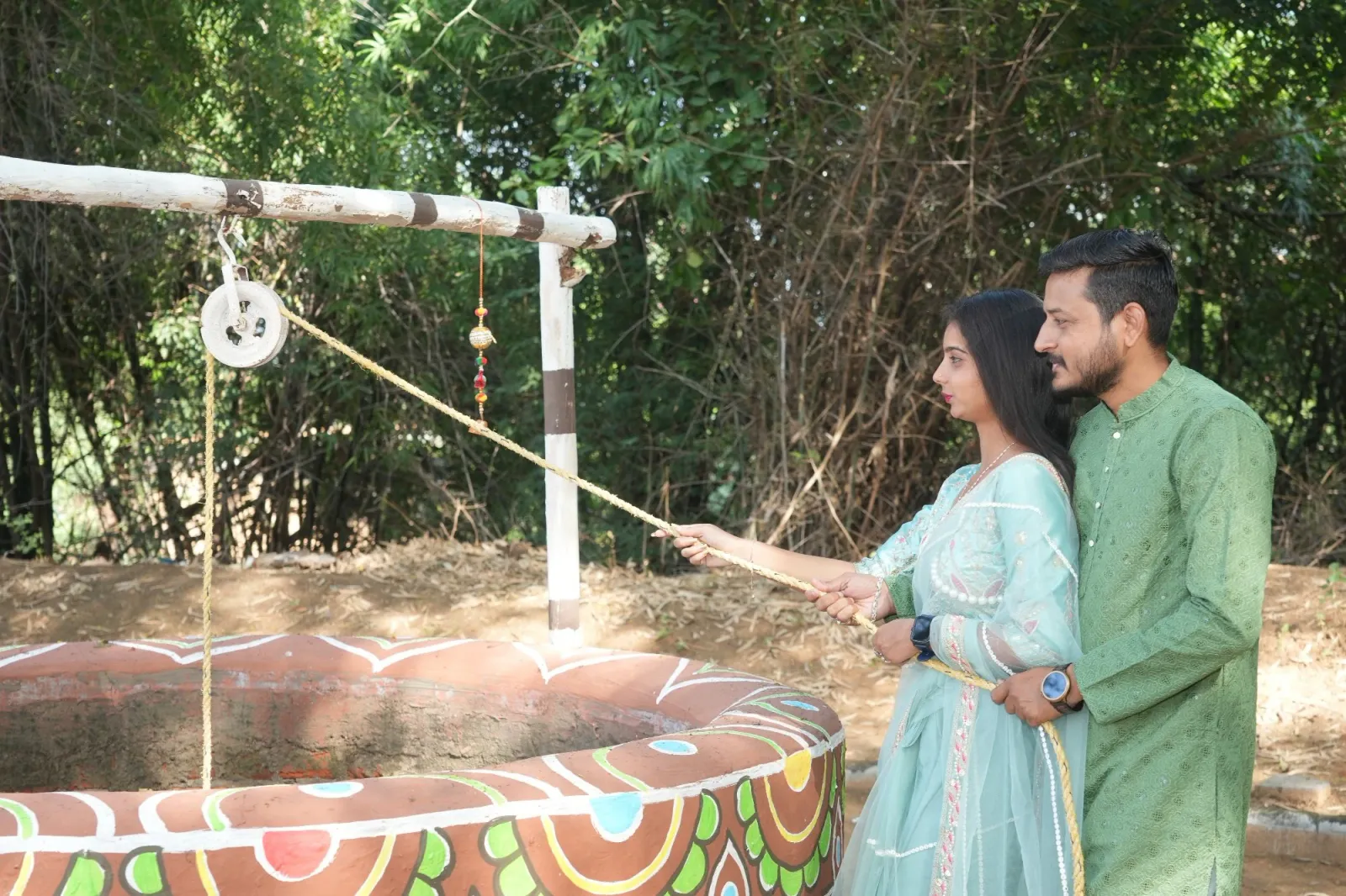 Couple in traditional wear draw water with a rope at a painted village well among green trees