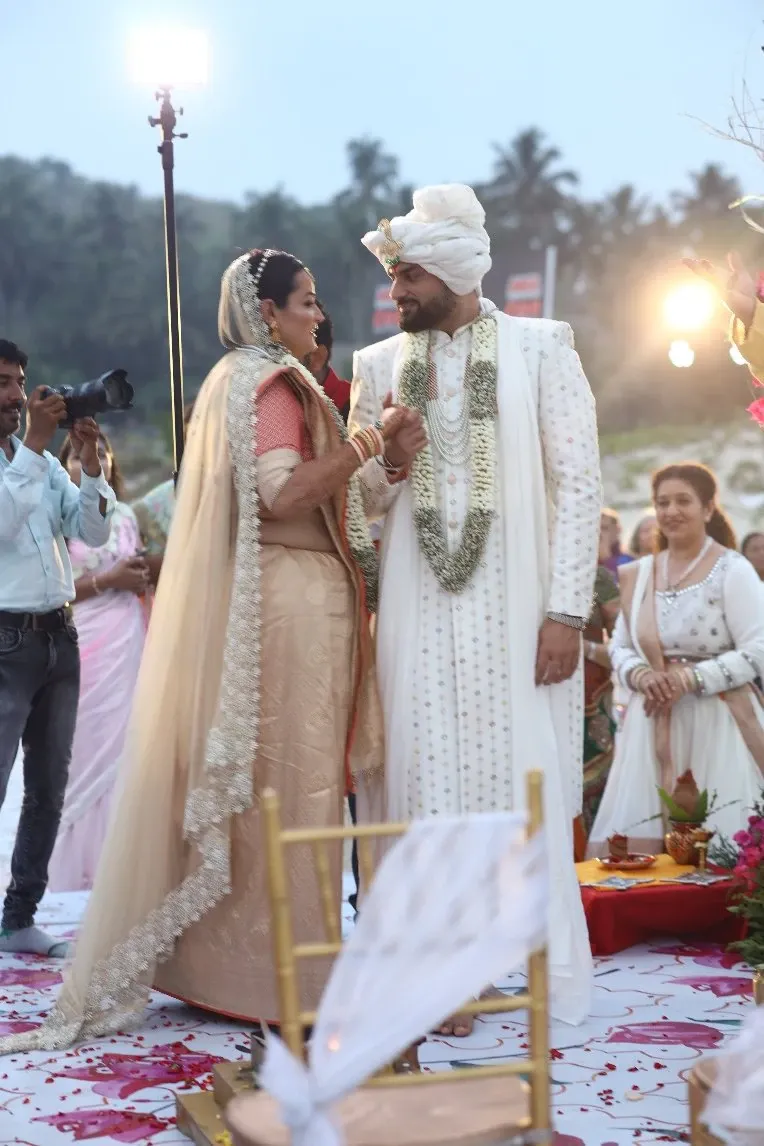 Bride and groom in wedding attire stand hand in hand on an outdoor mandap